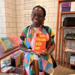 Mirella Maria stands holding a book titled: Souffles: On Deep Listening and Active Reception