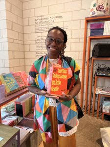 Mirella Maria stands holding a book titled: Souffles: On Deep Listening and Active Reception